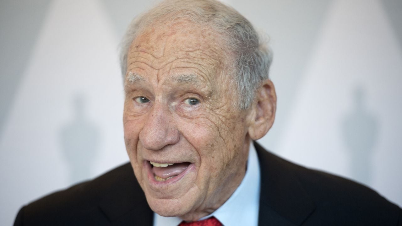 An older man with gray hair smiles widely while wearing a dark suit, white shirt, and red tie. The background is light with shadowy silhouettes visible behind him.