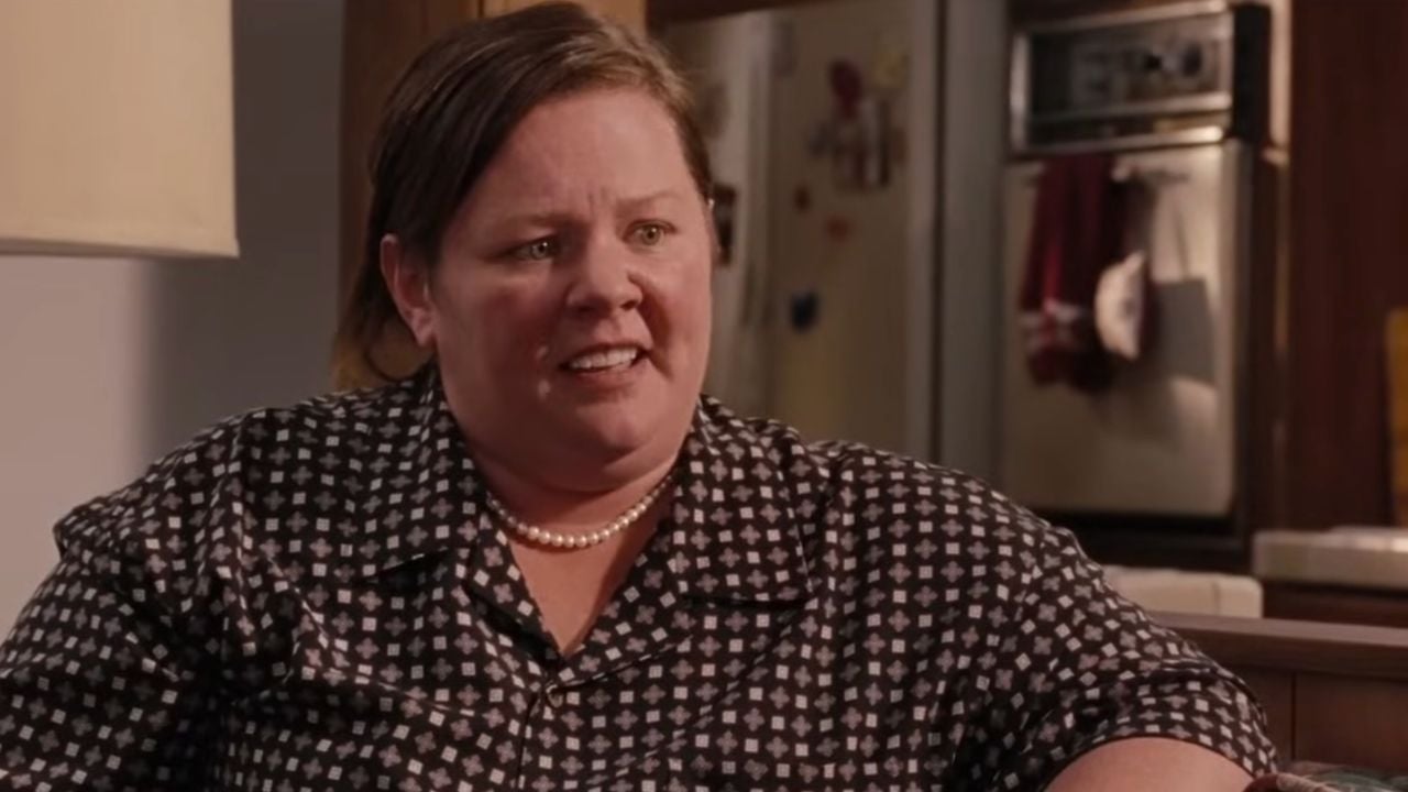 A woman with brown hair in a ponytail, wearing a black and white patterned blouse and pearls, sits indoors and appears to be talking, with a kitchen visible in the background.