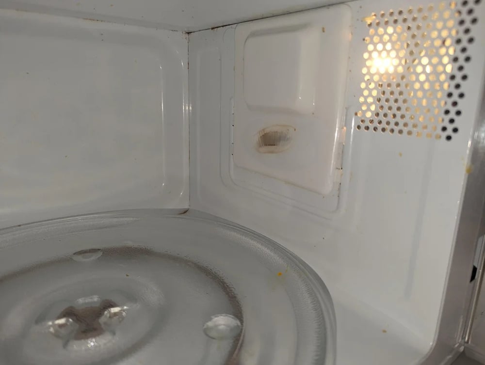 Close-up of the inside of a microwave oven, showing the glass turntable and a vent on the side, with minor stains and some grime visible near the vent opening.