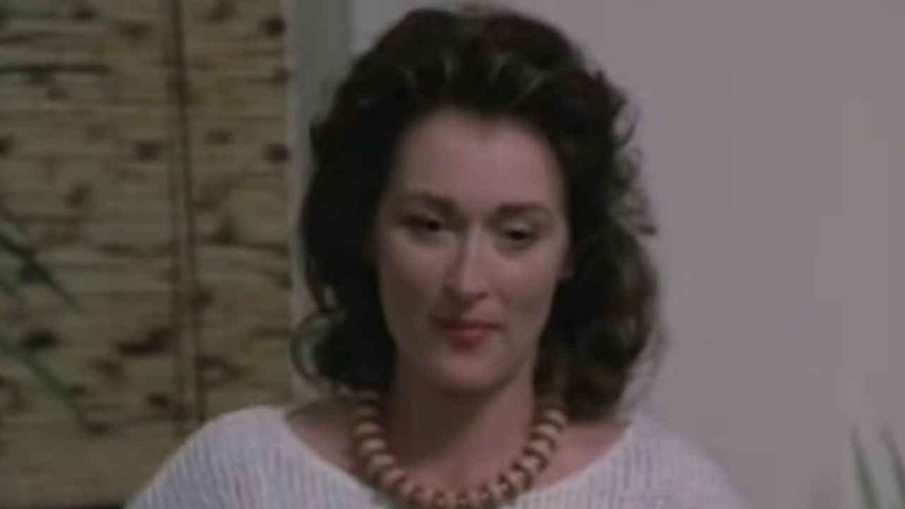 A woman with wavy dark hair wears a white top and a chunky wooden necklace, sitting indoors in soft lighting with a neutral expression.
