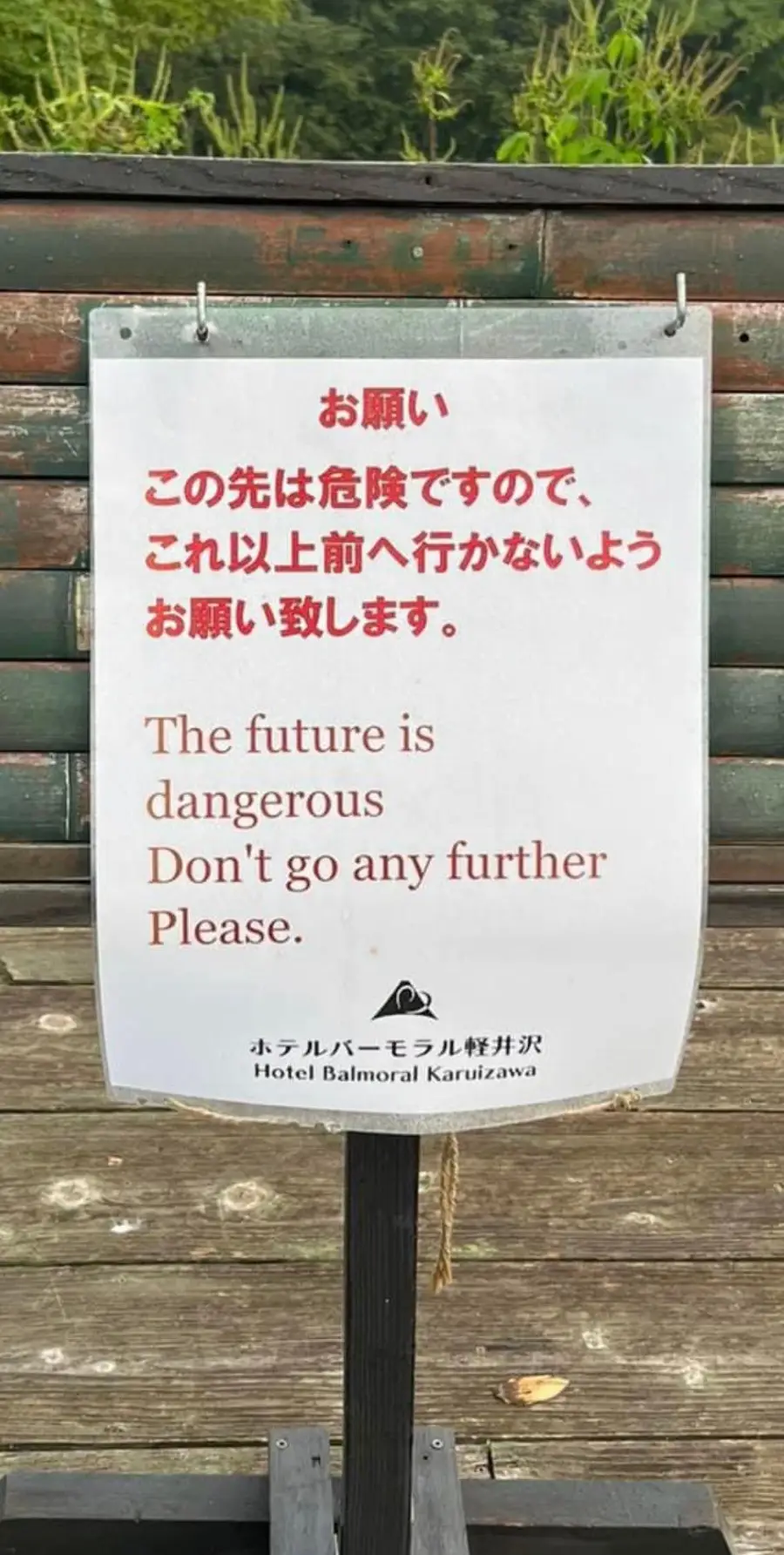 A sign in Japanese and English warns, “The future is dangerous. Don’t go any further. Please.” The sign is posted outdoors on a chain, with a wooden fence behind it.