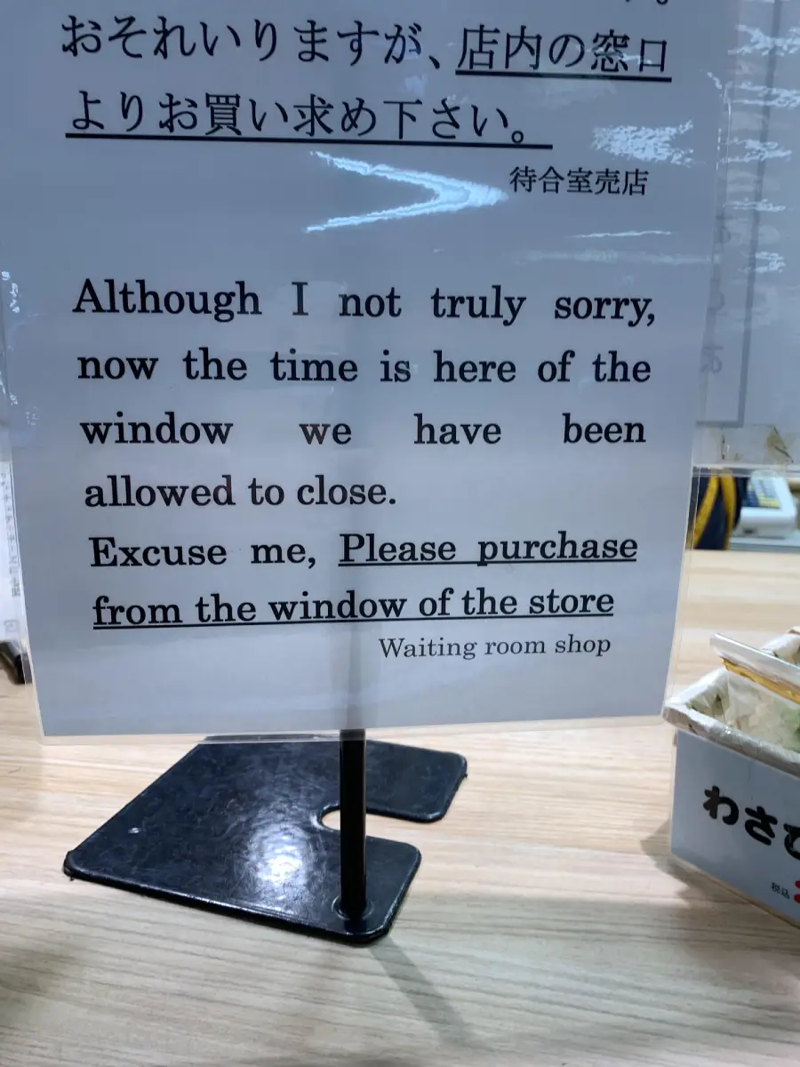 A sign in English asks customers to purchase from the window of the store, explaining that they can no longer buy inside. The text is formal and slightly awkward. The sign is on a counter with other signs nearby.