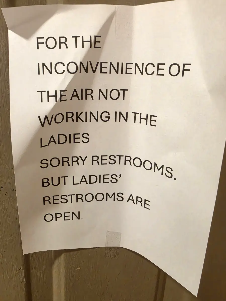 A printed sign apologizes for the air not working in the ladies restroom, but states that the ladies’ restrooms are open. The paper is taped to a surface and appears slightly wrinkled.