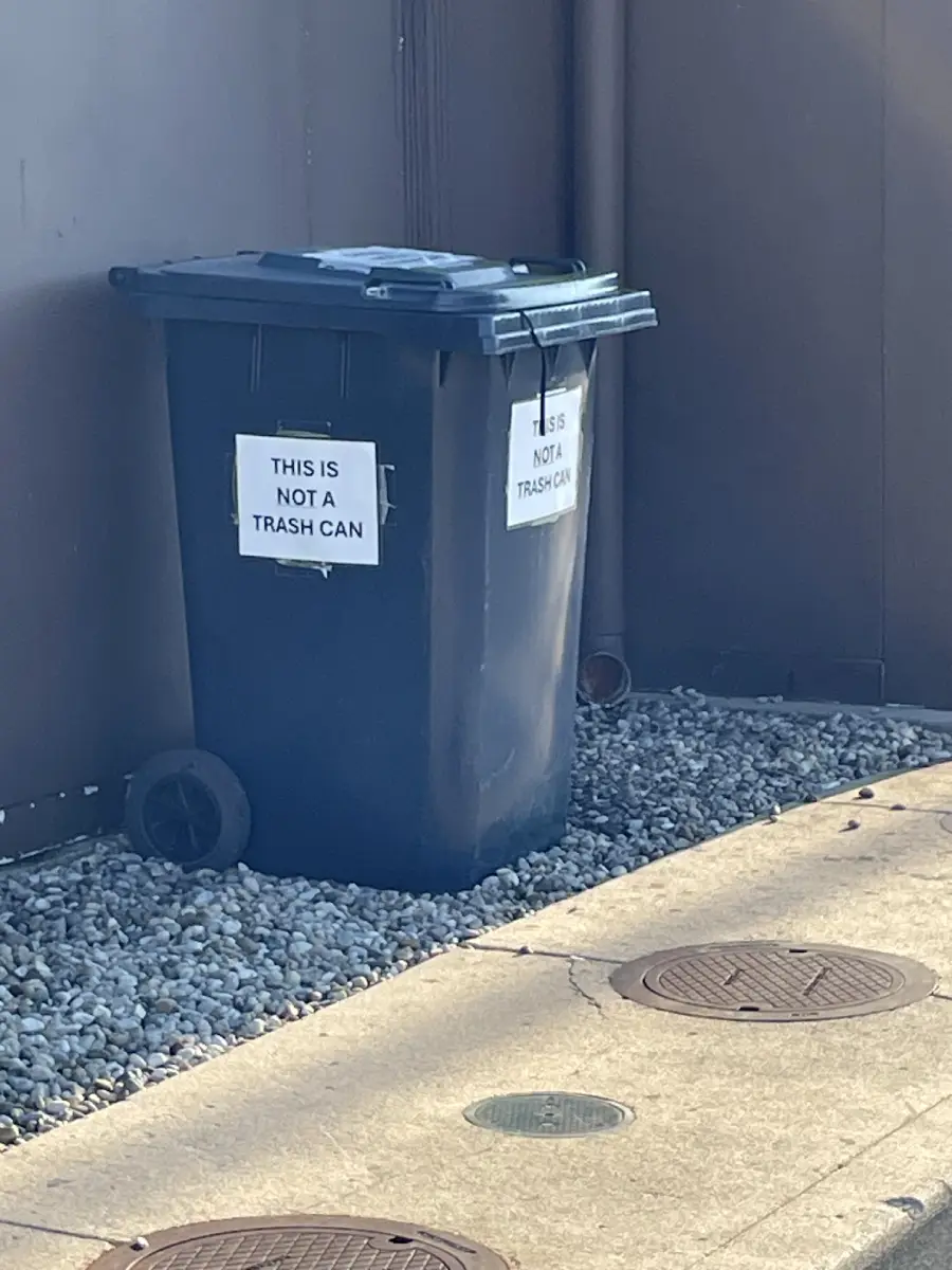 A black trash bin sits on gravel next to a wall, with two signs taped to it that both read, “THIS IS NOT A TRASH CAN.”