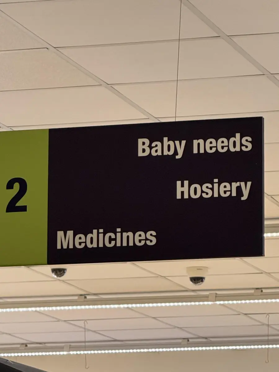 A store aisle sign labeled “2” lists “Baby needs,” “Hosiery,” and “Medicines” in white text on a black background, hanging from a ceiling with fluorescent lights and ceiling tiles visible above.