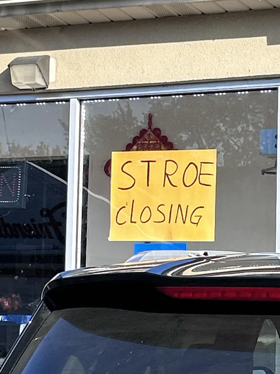 A handwritten yellow sign in a store window reads “STROE CLOSING” instead of “STORE CLOSING.” A black car is parked in front of the window.
