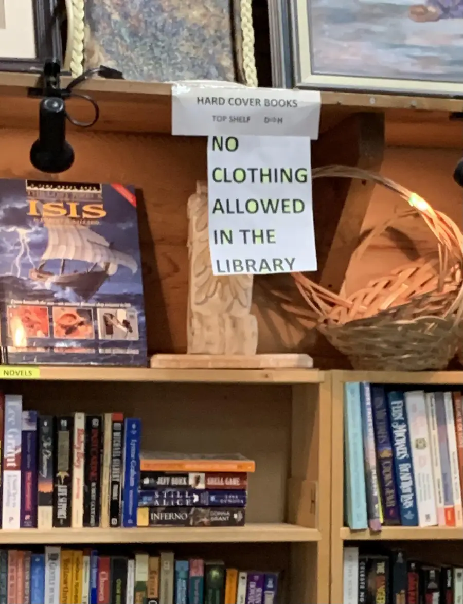A library shelf displays books and a sign that reads “NO CLOTHING ALLOWED IN THE LIBRARY.” Another sign above indicates “HARD COVER BOOKS TOP SHELF.” A wicker basket and statue are also visible.