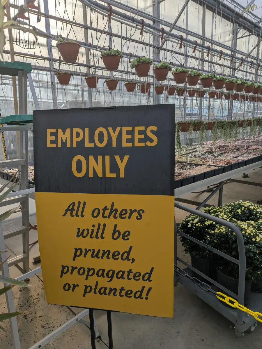 A sign in a greenhouse reads, “EMPLOYEES ONLY. All others will be pruned, propagated or planted!” Potted plants hang above, with flowers and plants on tables nearby.