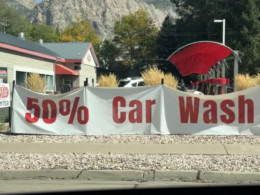 A large white banner with red text advertises “50% Car Wash” outside a car wash business, with mountains and trees visible in the background.