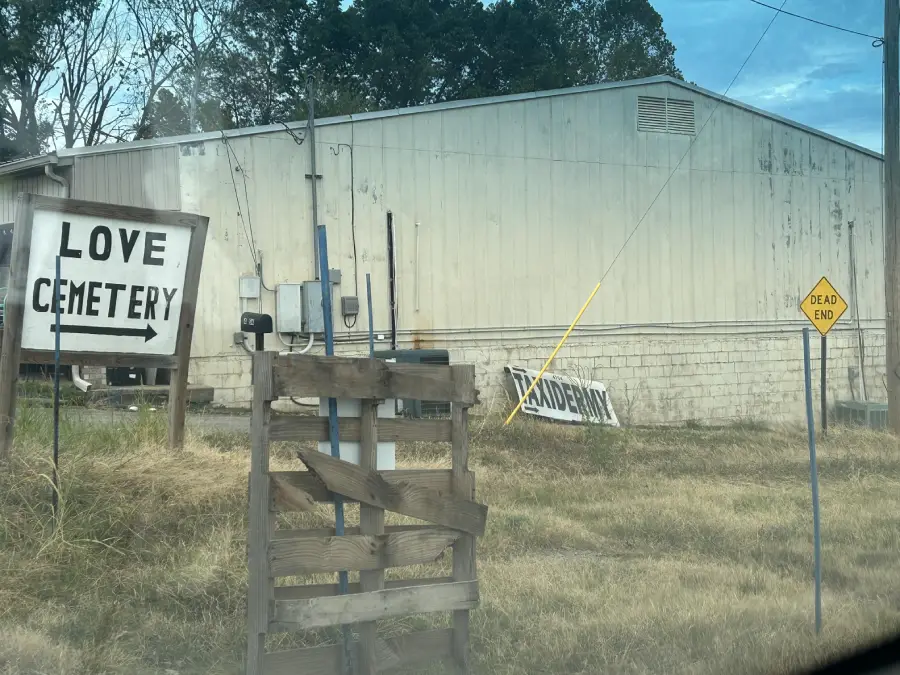 A weathered sign reads “LOVE CEMETERY” near an old building, with a tilted “DEAD END” road sign and a broken wooden fence in the foreground. Grass is overgrown and the sky is cloudy.