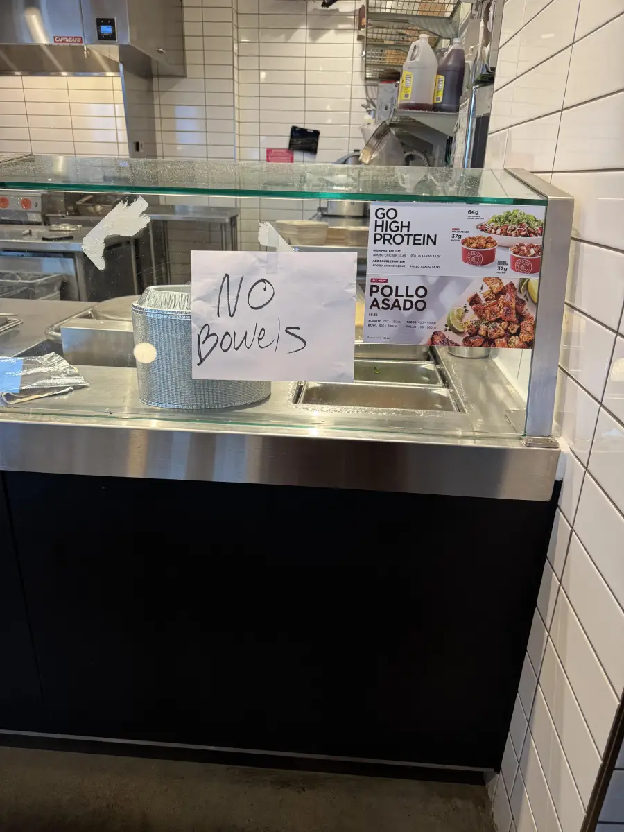A handwritten sign reading “NO Bowls” is taped to the glass above a counter at a fast food restaurant, next to a menu displaying high protein options and pollo asado.
