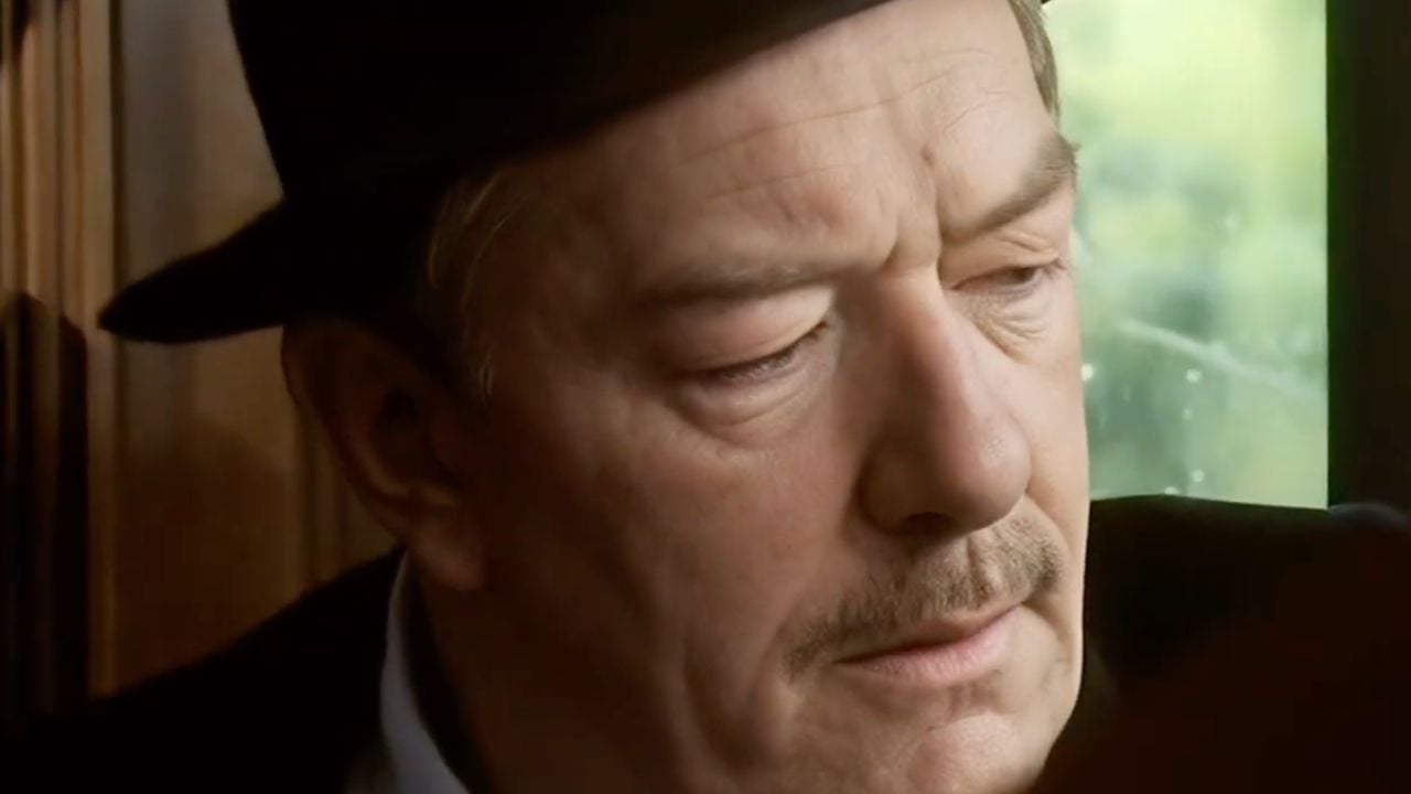 A man with a mustache and wearing a black hat looks down thoughtfully, seated by a window with green scenery visible outside.