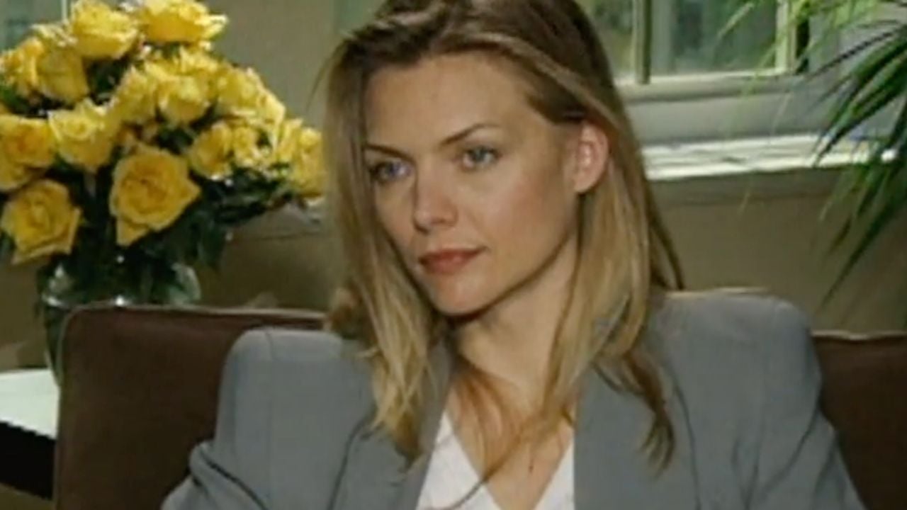 A woman with long blonde hair in a gray blazer sits indoors, yellow roses and greenery behind her, with a window in the background.