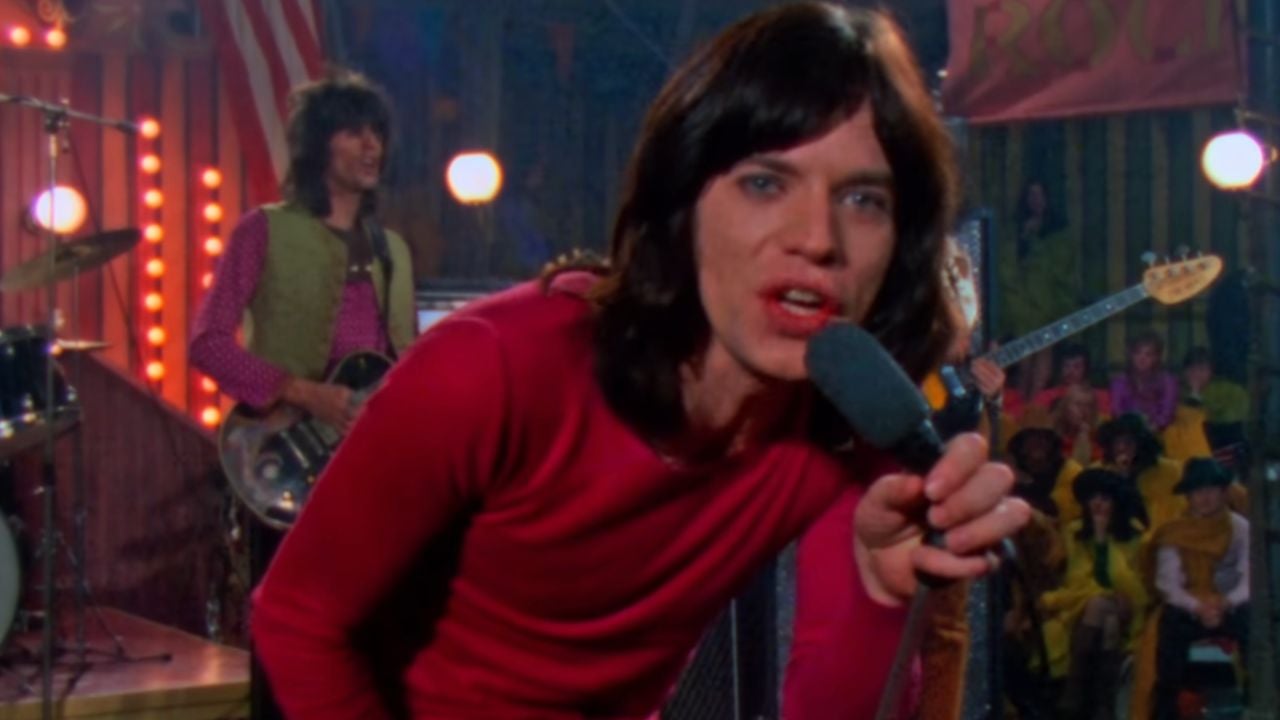 A man with shoulder-length dark hair in a red shirt sings into a microphone on stage, with a band and audience in the background.