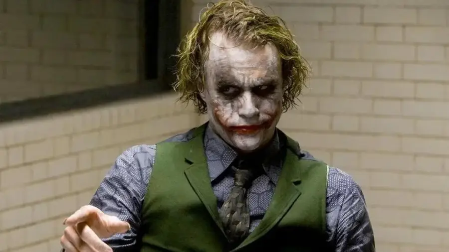 A person in white face paint with dark makeup, red lips, disheveled green hair, and wearing a green vest and tie stands in front of a tiled wall, gesturing with one hand.