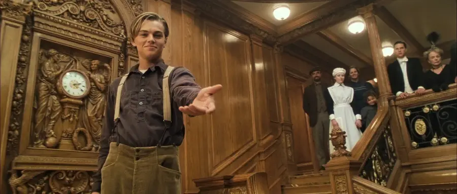 A young man stands on ornate wooden stairs, extending his hand forward. Behind him, several people watch from the landing. A decorative clock and carved designs are visible on the wall.