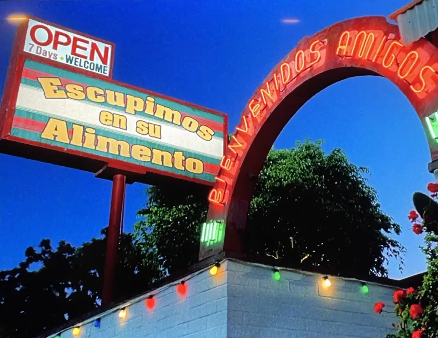 A restaurant sign reads “Escupimos en su Alimento” under an “OPEN” sign, with colorful lights and trees in the background. An arch says “Bienvenidos Amigos.”