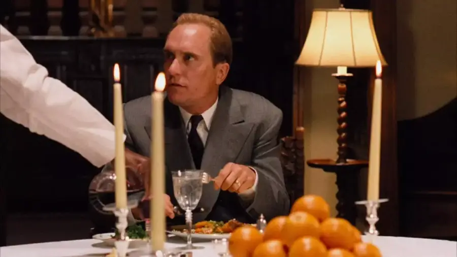 A man in a suit sits at a dinner table with candles, oranges, and a lamp, looking to his left as another person pours wine into his glass.