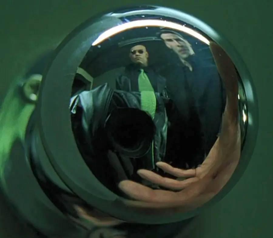 Two men are reflected in a shiny metal doorknob; one wears sunglasses and a green tie, the other wears black. A hand is partially visible at the bottom, with the scene tinted in green tones.
