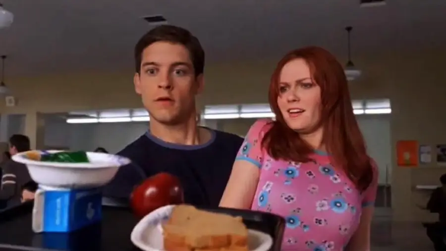 A young man in a blue shirt and a young woman in a pink floral shirt look surprised as a tray with food and a milk carton falls toward them in a cafeteria.