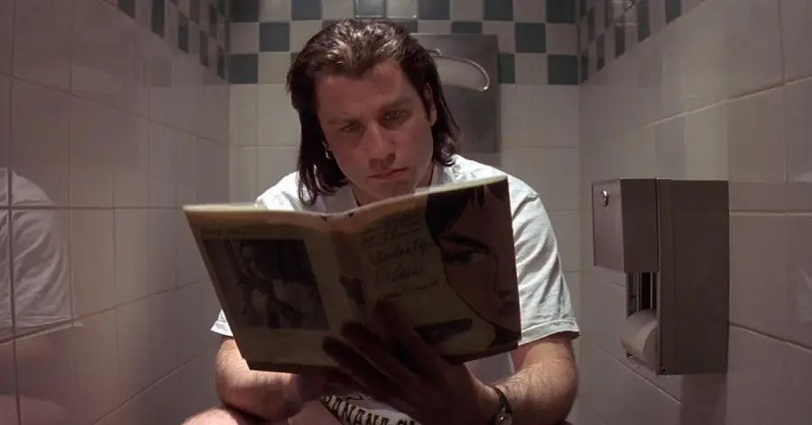 A man with long dark hair sits on a toilet in a tiled bathroom, reading a book with an illustrated cover. A metal toilet paper holder is mounted on the wall beside him.