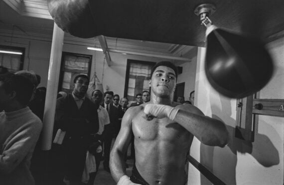 Muhammad Ali, shirtless and sweating, practices on a speed bag in a gym. He is surrounded by a crowd of onlookers watching his training session in this classic black-and-white photograph.