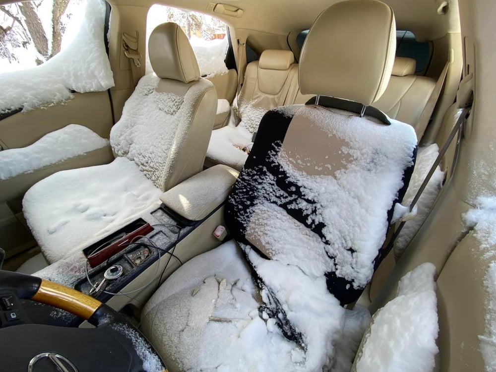 The interior of a car is covered in snow, with a thick layer on the seats, center console, and floor, suggesting the windows or doors were left open during a snowfall.
