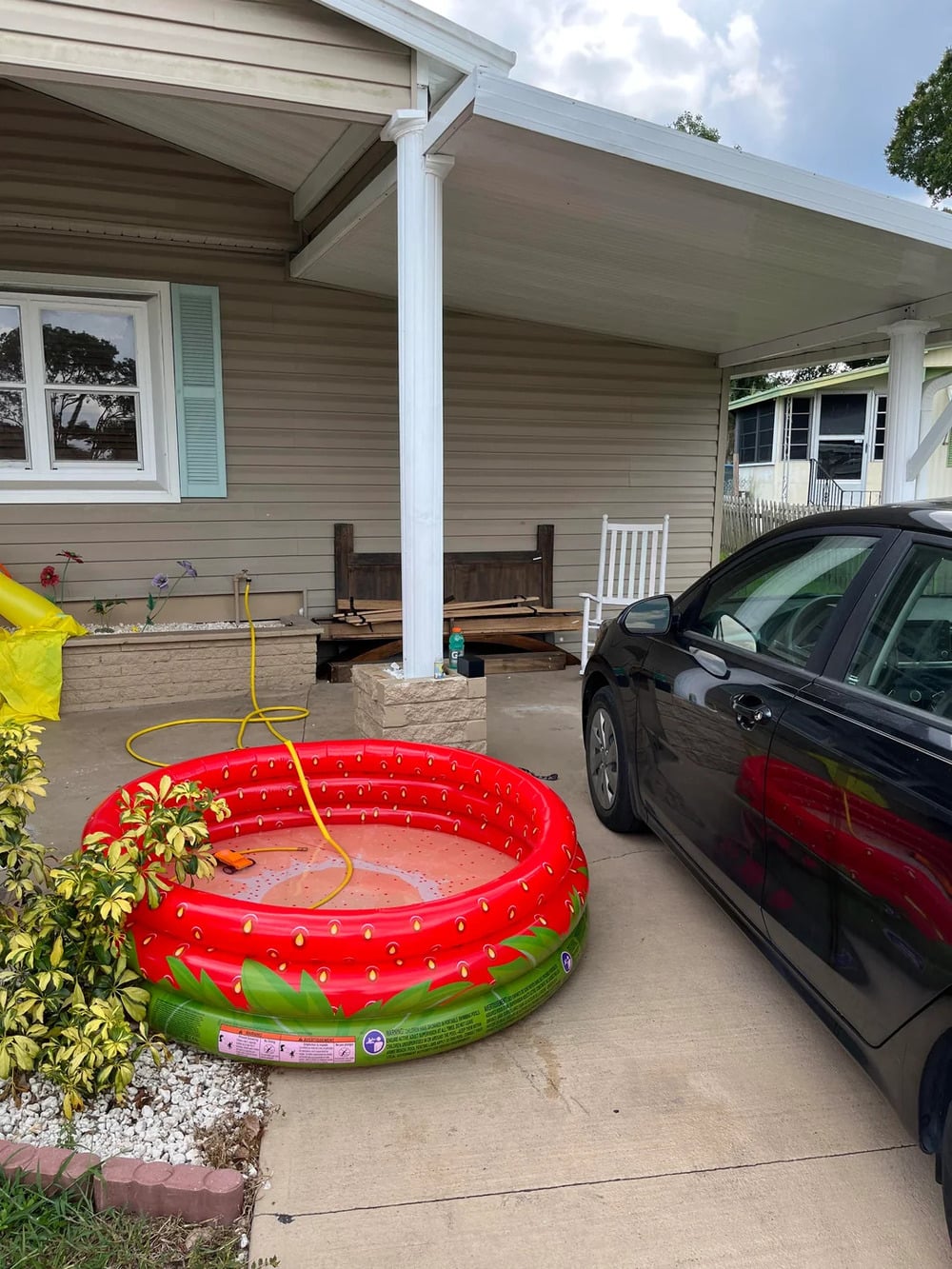 A small, red inflatable pool filled with water sits on a driveway near a black car in front of a beige house with a covered porch and a white rocking chair.