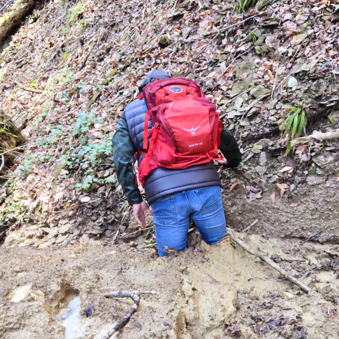 A person wearing a red backpack and blue jeans is walking through muddy terrain in a forest, with their back facing the camera. The ground is wet and slippery, surrounded by fallen leaves and vegetation.