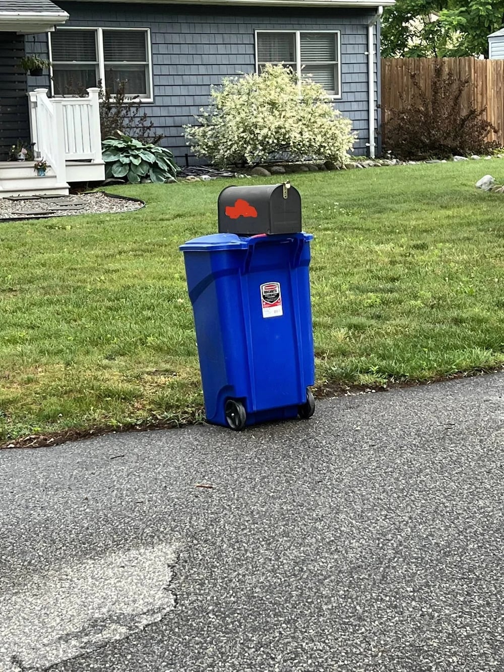 A blue recycling bin is placed on the edge of a driveway, directly beneath a black mailbox with a red flag, in front of a gray house with a porch and green lawn.