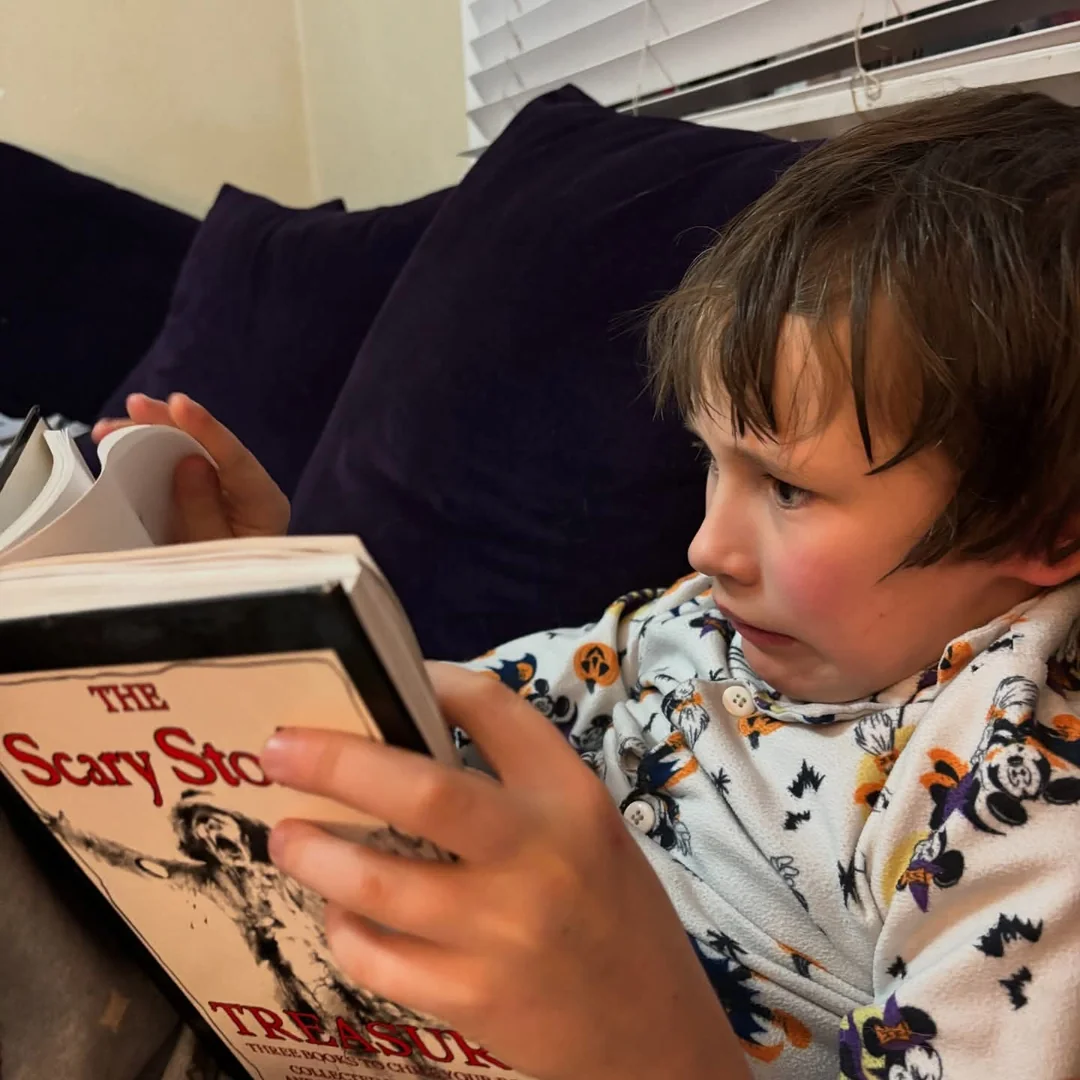 A child in Halloween-themed pajamas sits on a couch reading “The Scary Story Treasure” book, looking focused and slightly concerned.