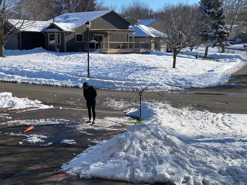 A person in dark clothes stands near a pile of snow on a driveway, with snow shoveled to the side. A shovel is stuck upright in the snow, and a carrot lies on the ground. Houses and trees are in the background.