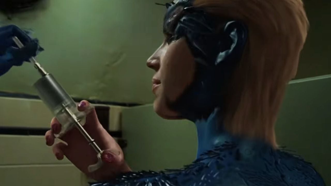 A person with blue skin and blonde hair holds a large syringe while looking at it closely in a dimly lit room.