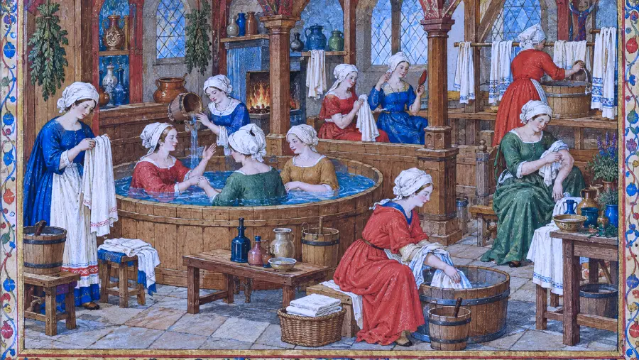 Medieval women in dresses and white caps bathe, chat, and wash laundry in a wooden bathhouse with stained glass windows, wooden tubs, and shelves lined with towels and jars.