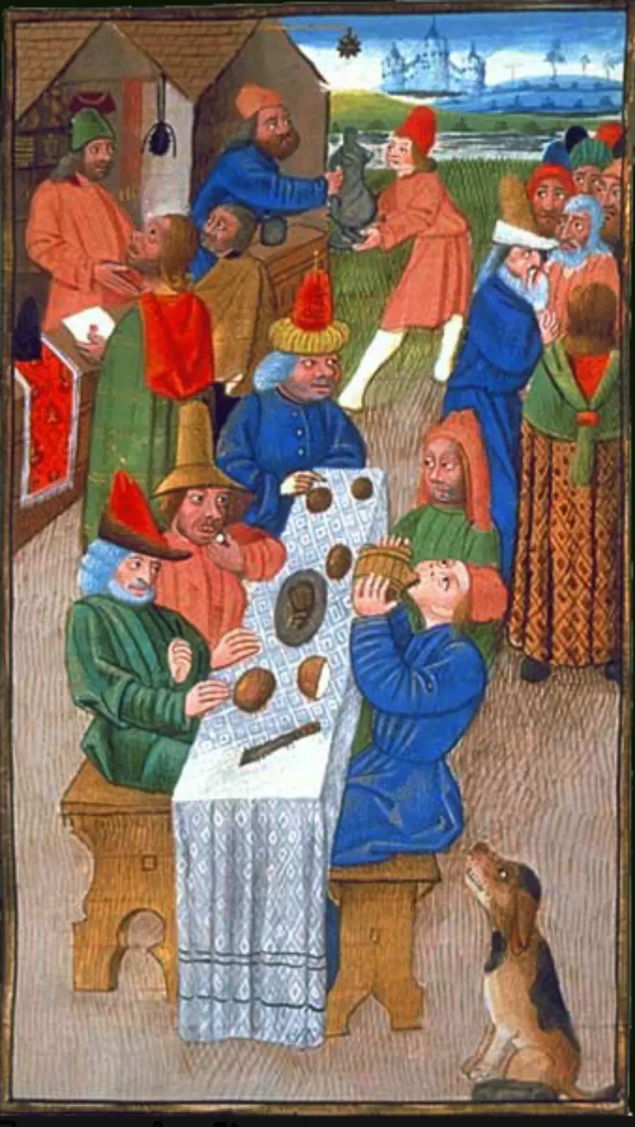 Medieval illustration of people in colorful robes feasting at a long table outdoors, some eating and drinking, others talking or serving food, with a dog sitting by the table and a village in the background.