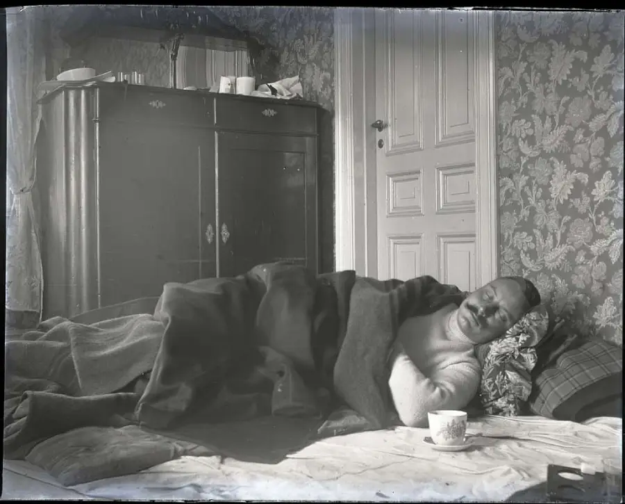 A man lies in bed under heavy blankets, resting his head on a pillow. A teacup sits on the blanket in front of him. Behind him are a wardrobe, a door, and patterned wallpaper.