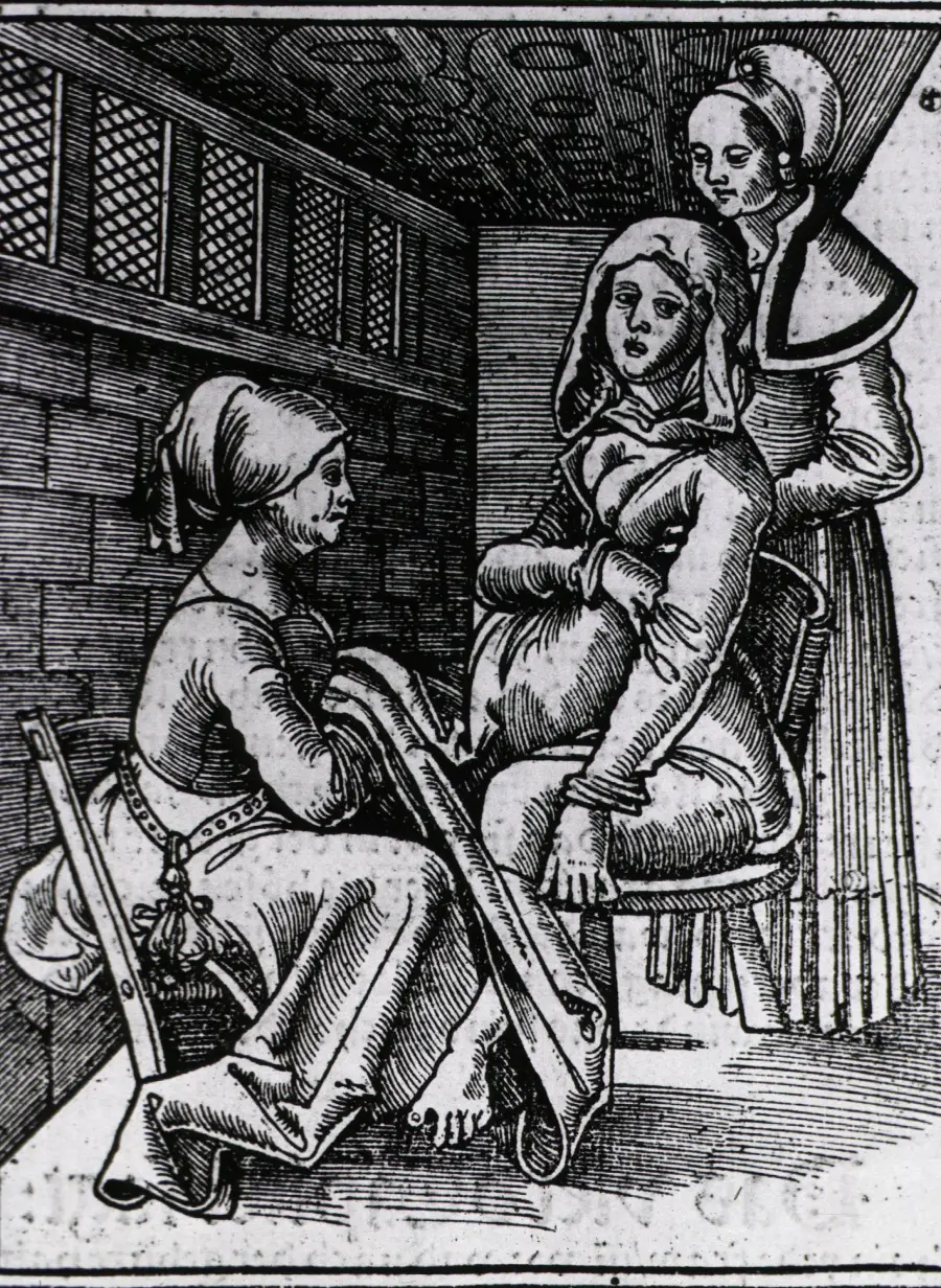 A black and white woodcut shows a woman in labor seated, assisted by two women in old-fashioned clothing inside a room with brick walls, one kneeling, holding the mother’s legs.