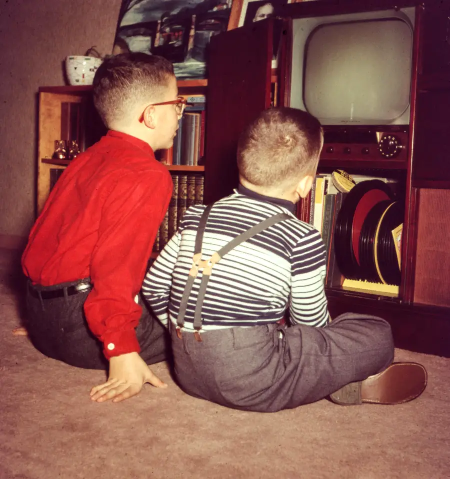 Two young boys sit on the floor facing a vintage television set. One wears a red shirt and glasses, the other wears a striped shirt with suspenders. Records are visible on a shelf beside the TV.