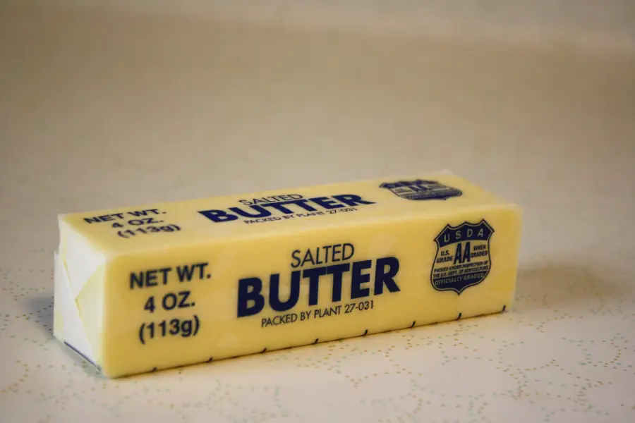 A stick of salted butter in its wrapper, labeled 4 oz. (113g), sits on a light surface. The packaging displays “SALT BUTTER” in blue text along with USDA AA certification.