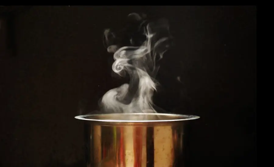 A metal pot on a dark background with white steam rising in delicate swirls from the top, suggesting something hot is being cooked or heated inside.