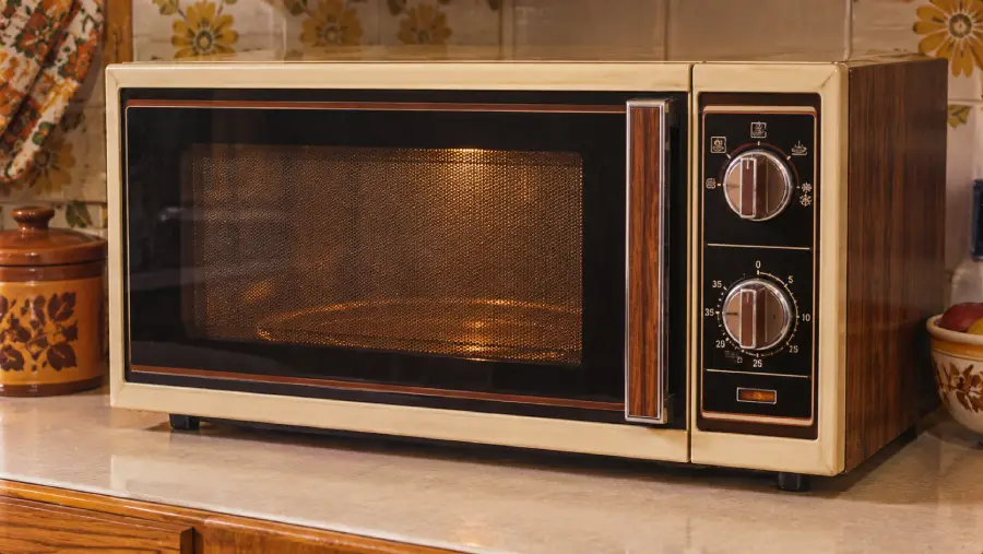 A vintage microwave oven with dials is on a kitchen counter. The background features a floral patterned backsplash and various kitchen items, including a ceramic jar and a bowl of fruit.