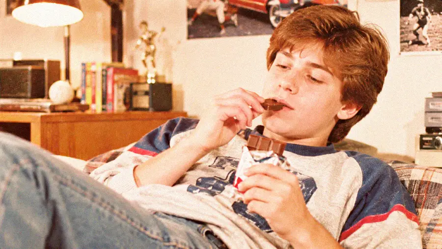A teenage boy in casual clothes lounges on a couch, eating a chocolate bar and looking relaxed. The background shows books, a trophy, and posters on the wall.