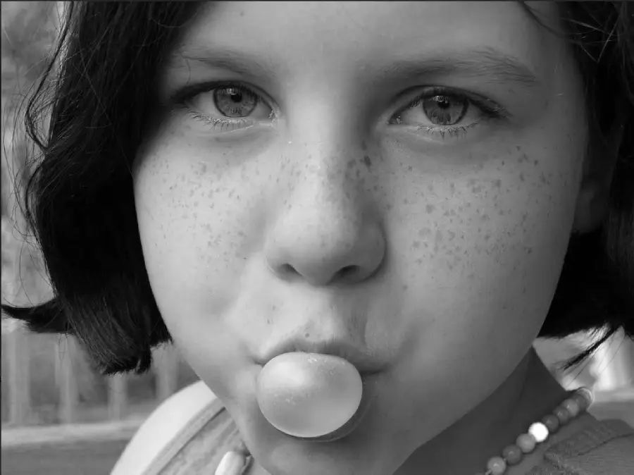 A close-up of a young person with short dark hair and freckles blowing a small bubble with gum. The photo is in black and white, and the person is wearing a beaded necklace.