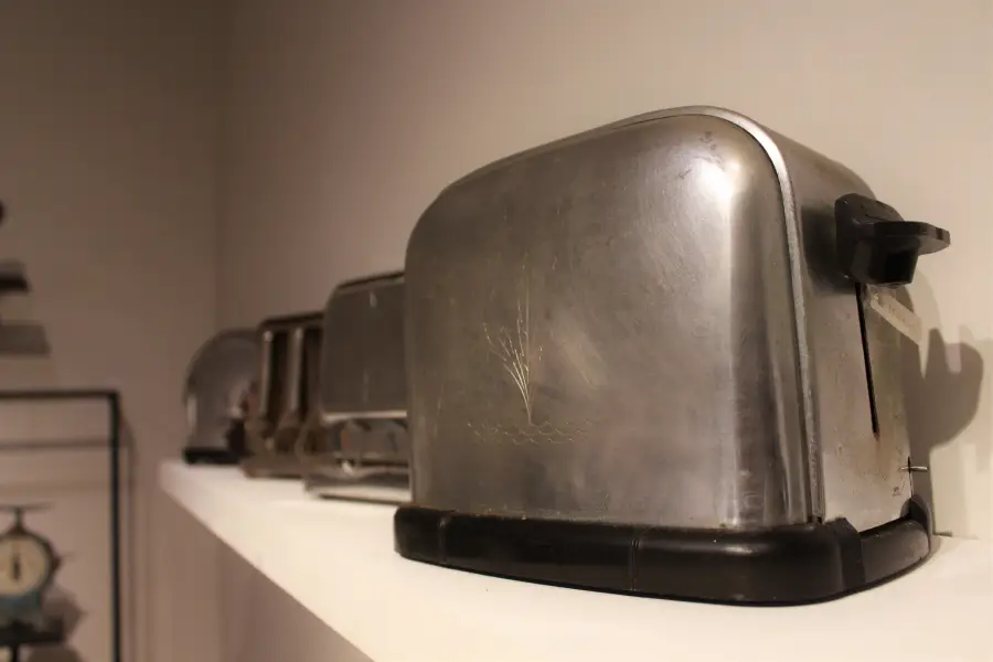 A row of vintage metal toasters sits on a white shelf against a plain wall, with the focus on the nearest silver toaster.