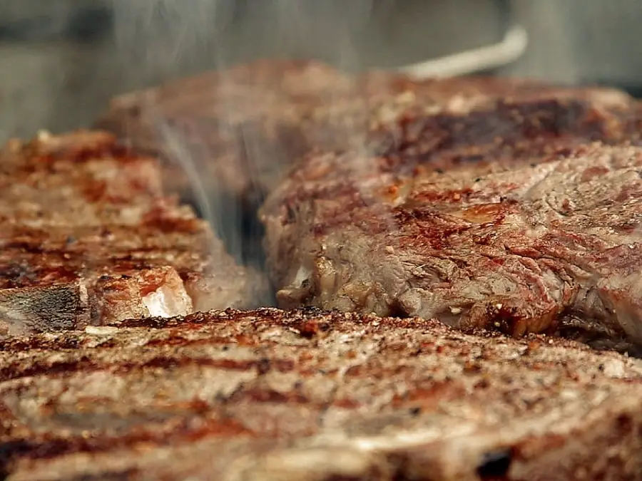 Close-up of several thick, juicy steaks sizzling and smoking on a grill, showing a browned, seared crust and hints of grill marks, with steam rising from the meat.