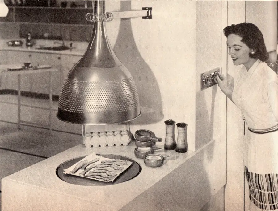 A woman in a 1950s kitchen smiles while turning a wall dial. Bacon is cooking under a large, futuristic hood on a counter, with eggs and condiments nearby. The scene has a retro, technological feel.