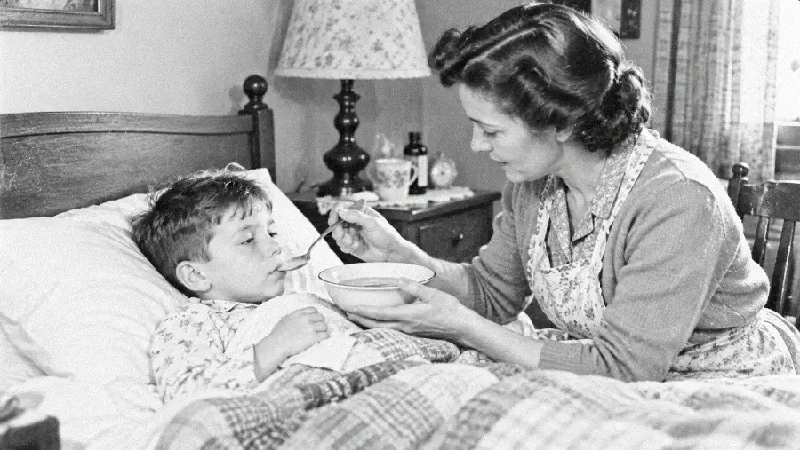 A woman sits beside a bed, feeding soup to a young boy who is lying under a blanket. The scene appears to be from the past, with vintage decor and clothing visible in the room.