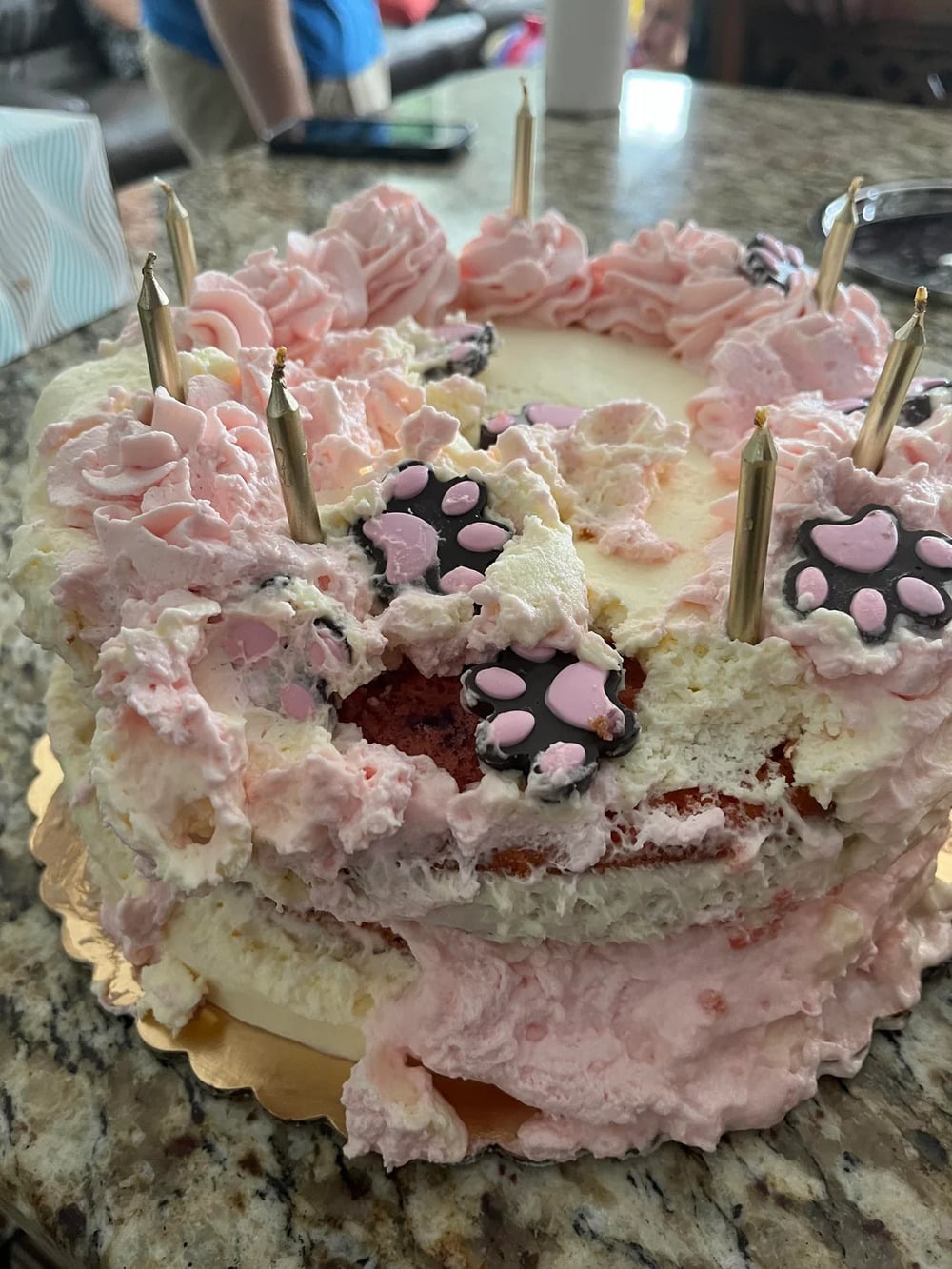 A partially destroyed pink and white cake with candles and paw print decorations sits on a granite counter. Some frosting is smeared and pieces are missing.