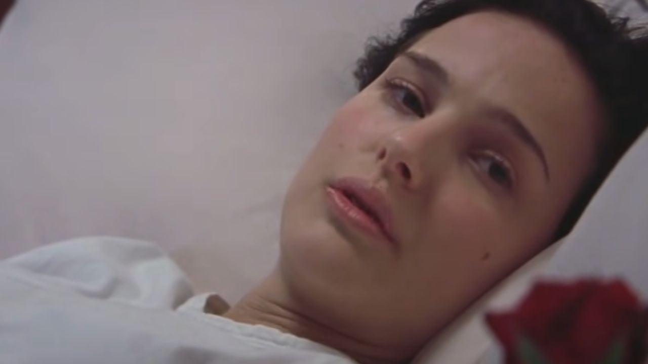 A woman with short dark hair lies on a white pillow, looking tired or unwell. She is wearing a white top and gazing softly ahead. A red rose is visible in the foreground.