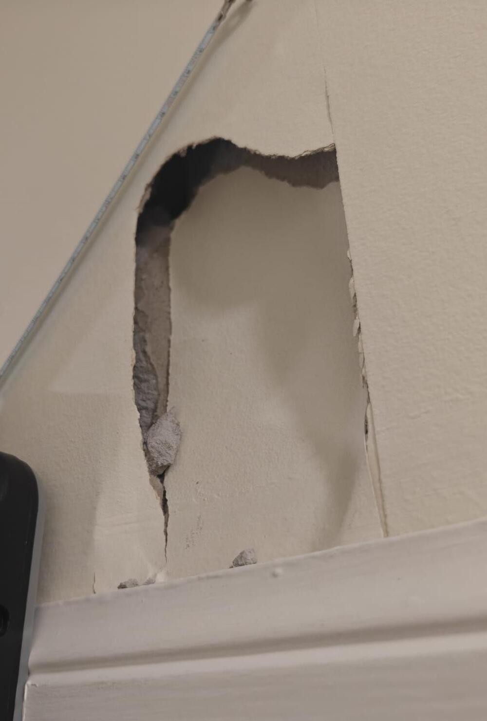 A close-up of a wall with a large hole exposing drywall and insulation above white trim, with a black object partially visible in the lower left corner.